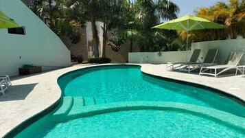 Une piscine à bord incurvé, entourée de chaises longues blanches et d’un parasol vert.