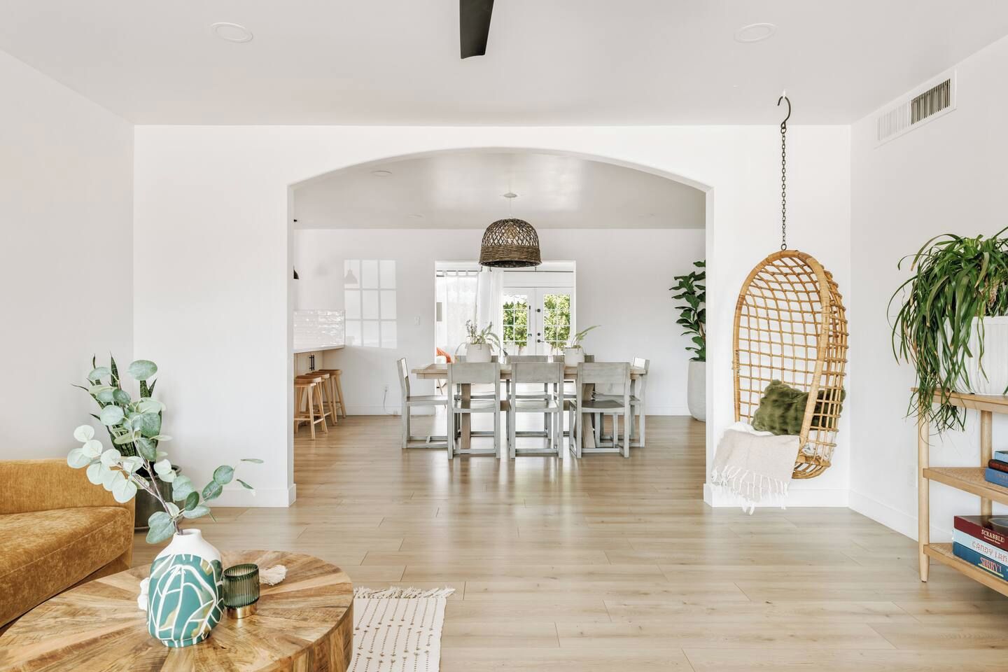 A spacious living room with a wooden floor, a sofa, a coffee table with decorative items, a hanging chair, and a view into the dining area with a table and chairs.