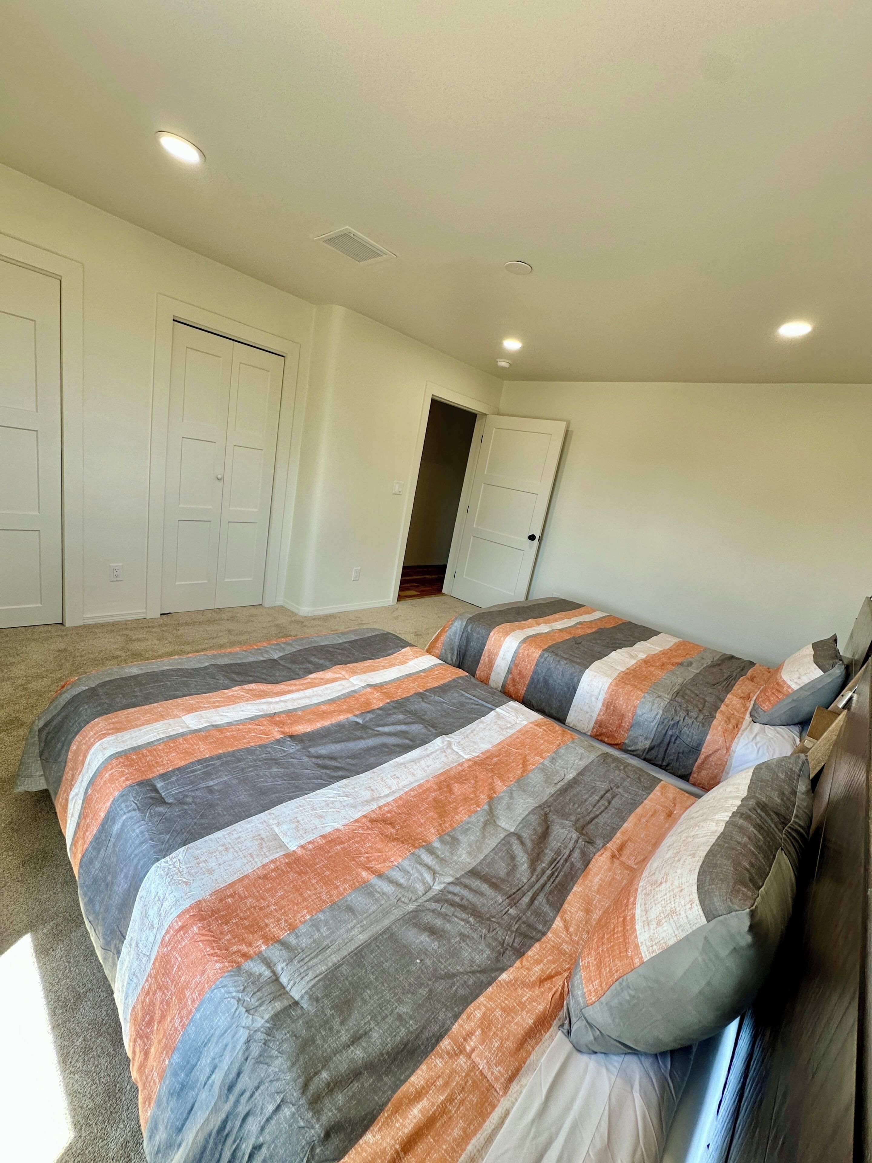 Un dormitorio con dos camas, piso alfombrado, paredes blancas y una puerta que da a otra habitación.