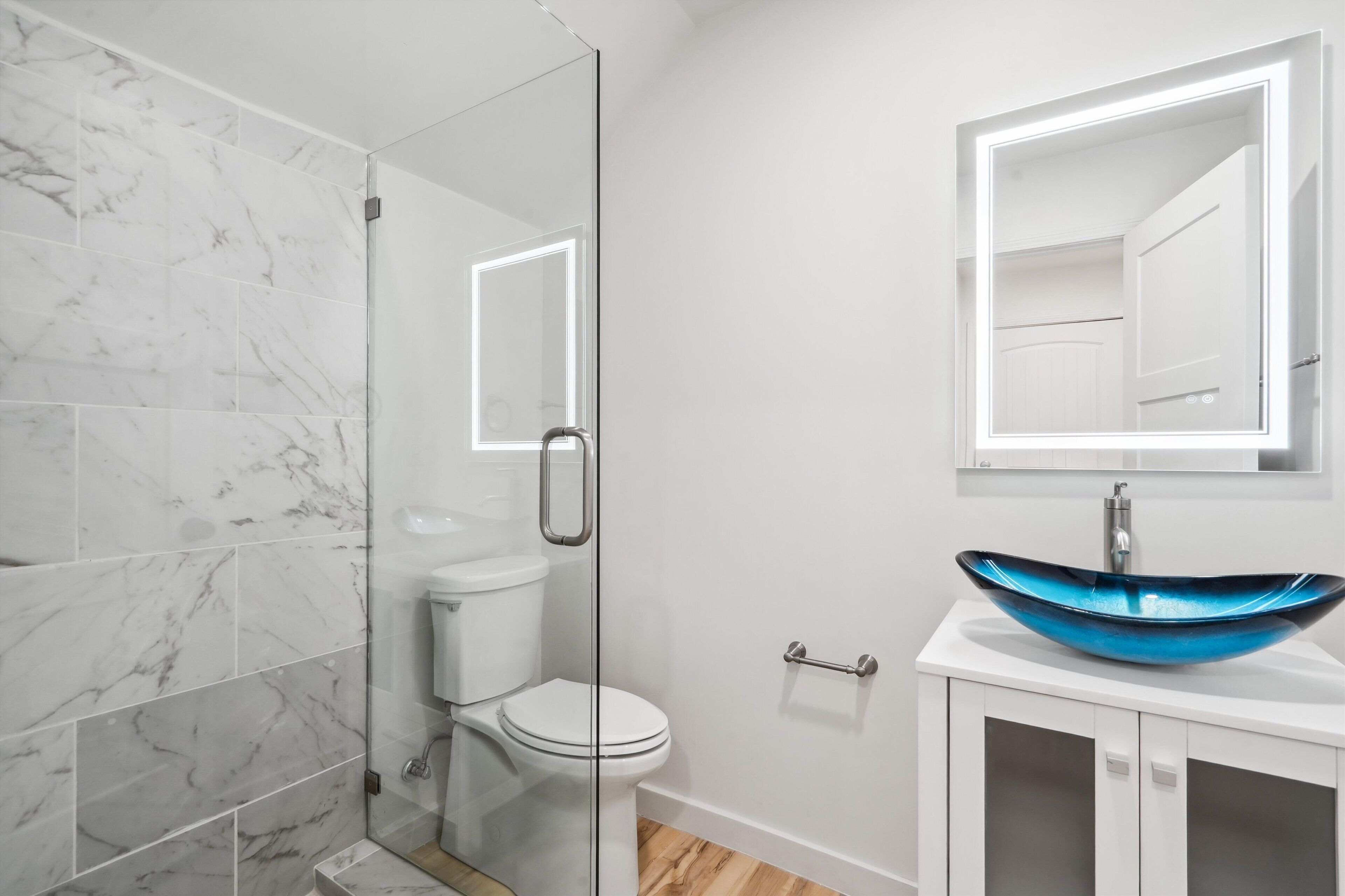 Une salle de bain dotée d’une douche à parois de verre, de toilettes blanches, d’un lavabo bleu et d’un miroir avec éclairage intégré.