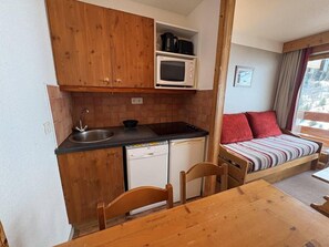 A compact kitchen with wooden cabinets, a microwave, a sink, and a dishwasher. There is a small dining table with chairs. A bed with a striped blanket and red pillows is visible in the background. - 300m from resort center, 9th floor, balcony, ski locker, 26m², Méribel-Mottaret (Méribel - Les Allues)