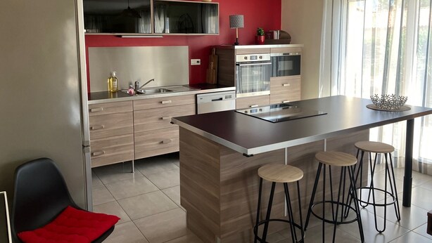 A modern kitchen with a central island, bar stools, a red accent wall, and a large window with curtains. - Maison Avalone in the heart of the Ardèche Gorges! (Vallon-Pont-d'Arc)