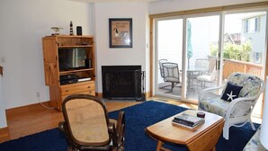 A living room with a fireplace, a television, a bookshelf, and a view of the backyard through a sliding glass door. - Tori's Landing- Limited Water View Shared Pier Access (Chincoteague)