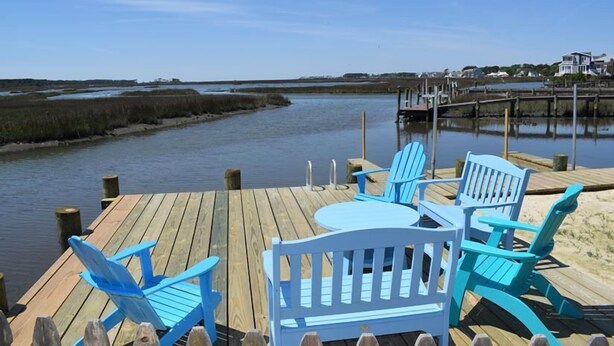 A wooden deck with blue Adirondack chairs and a small table, overlooking a calm body of water with a dock in the background. - Tori's Landing- Limited Water View Shared Pier Access (Chincoteague)