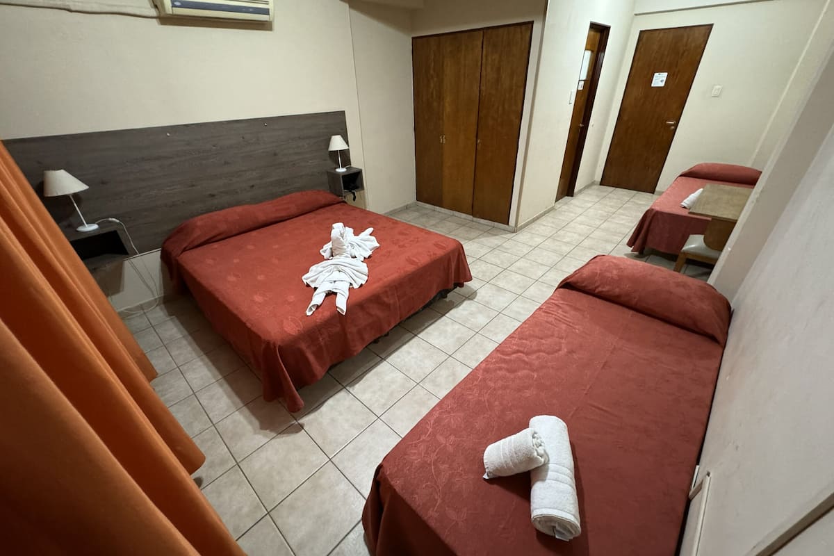 A hotel room with two beds, a wooden headboard, a bedside table with a lamp, and a bathroom with a shower and toilet.