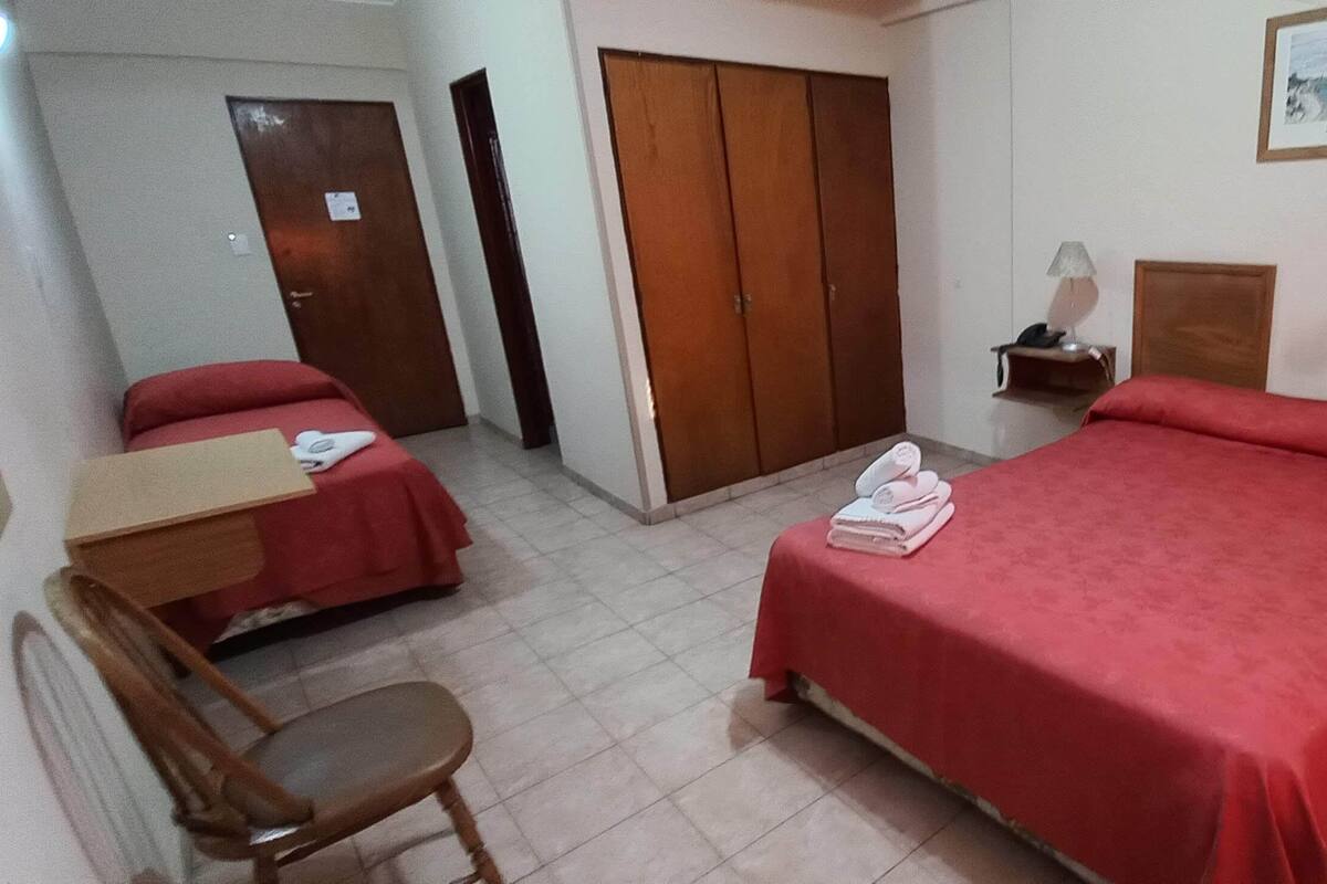 A hotel room with two single beds, a desk, a chair, a wardrobe, and a bedside table with a lamp.