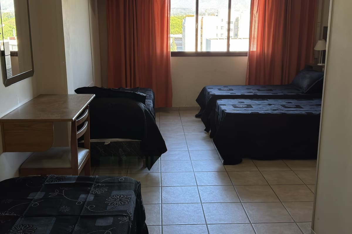 A hotel room with a desk, two beds, a window with curtains, and a view of buildings.