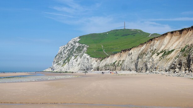 A beach with a prominent white cliff and a green hill in the background.