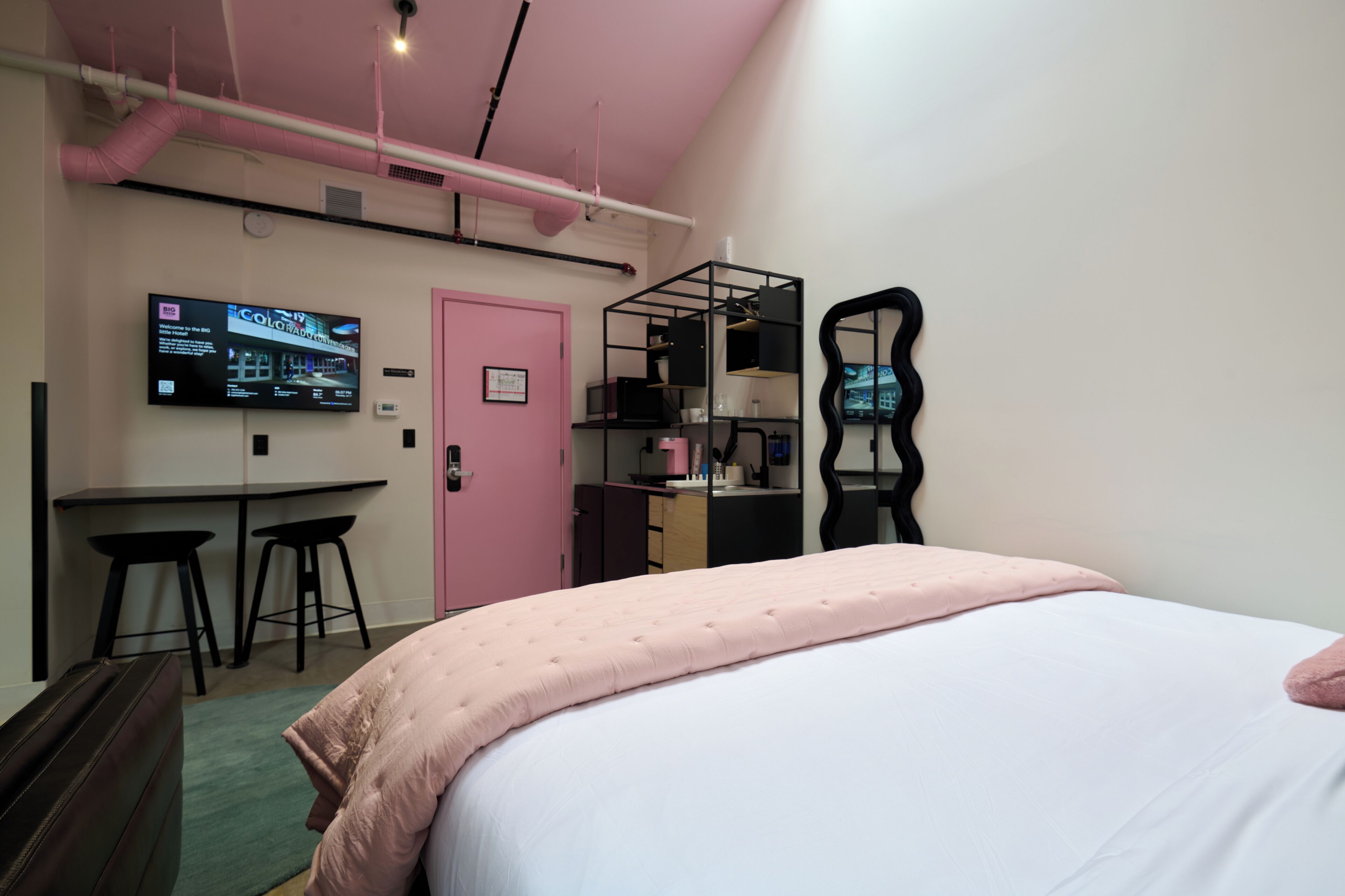 A bedroom with a bed, a desk with two stools, a pink door, and a TV on the wall.