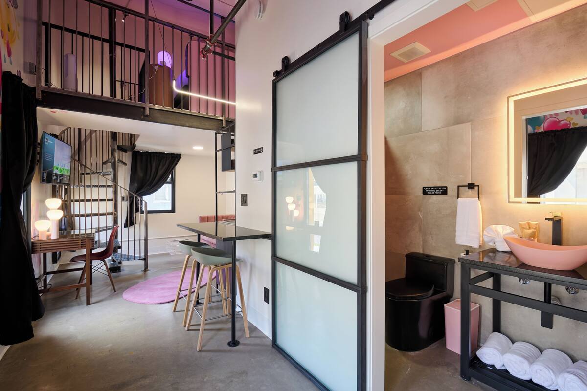 Habitación de hotel compacta con cama en literas, un pequeño comedor, y un baño con un espejo grande y un lavabo rosa.