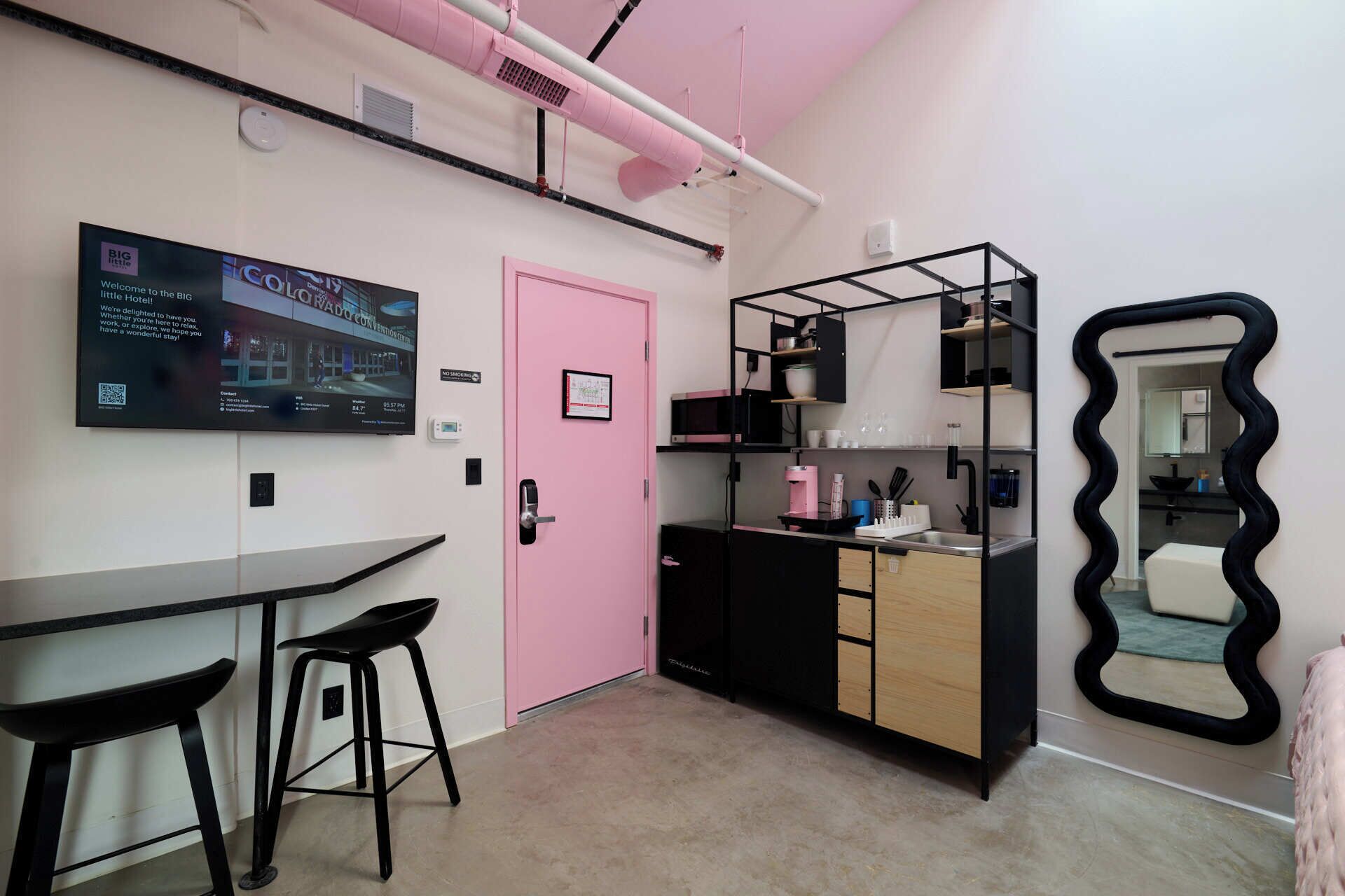 A modern kitchen with a pink door, black and white cabinets, and a black countertop. There is a bar stool and a large mirror with a wavy frame.