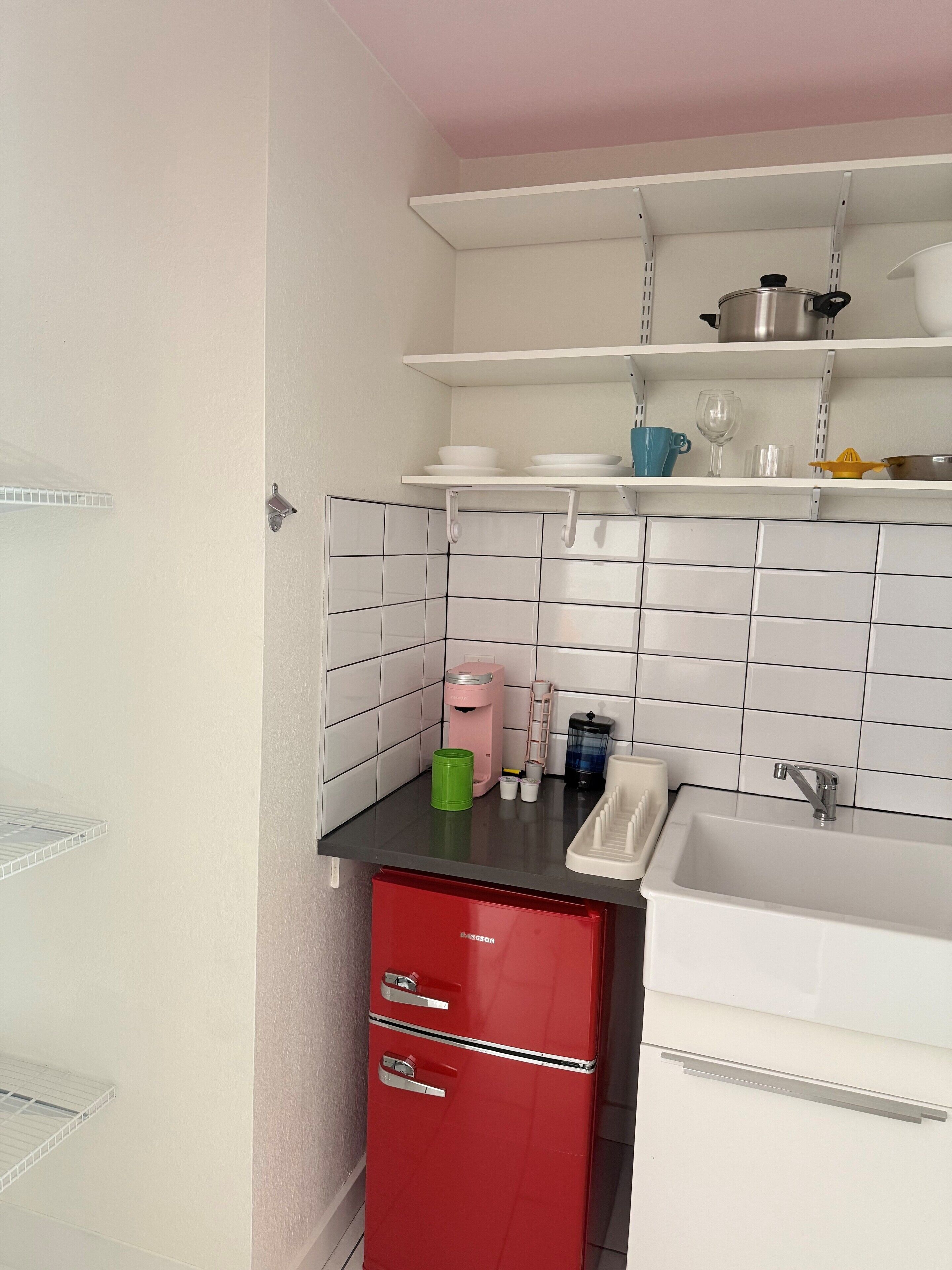 Una cocina compacta con salpicadero de azulejos blancos, un lavabo, un armario rojo y una encimera con diversos utensilios de cocina.