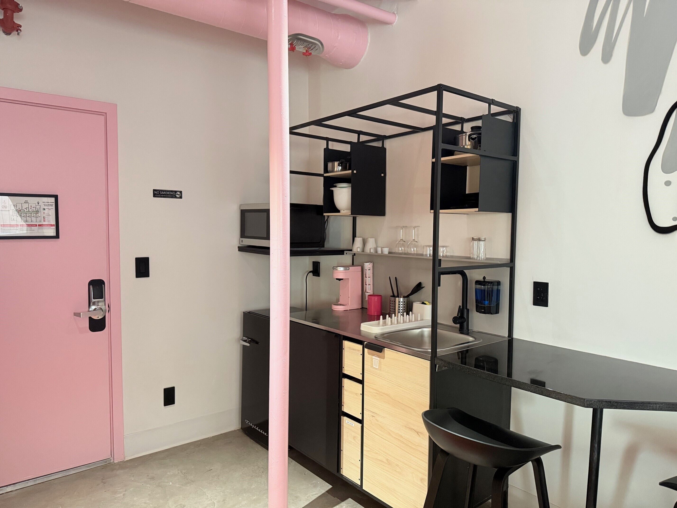 Una kitchenette con una puerta rosa, un estante metálico negro y una encimera negra con un lavamanos.