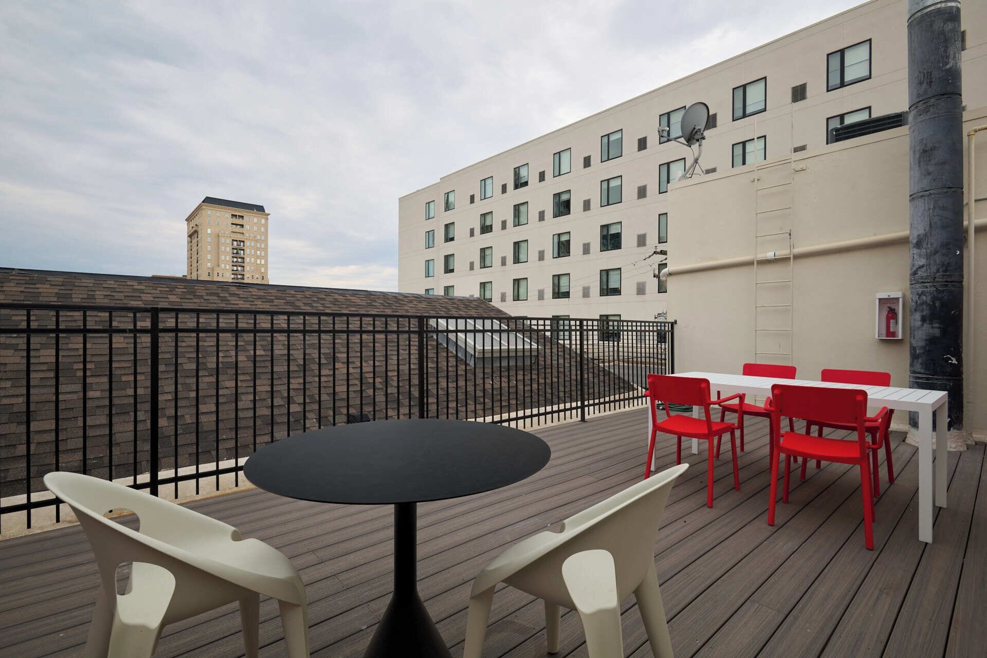 Une terrasse sur le toit, avec une table ronde et des chaises rouges et blanches, d’où l’on peut admirer un immeuble moderne doté de nombreuses fenêtres et d’une antenne parabolique.
