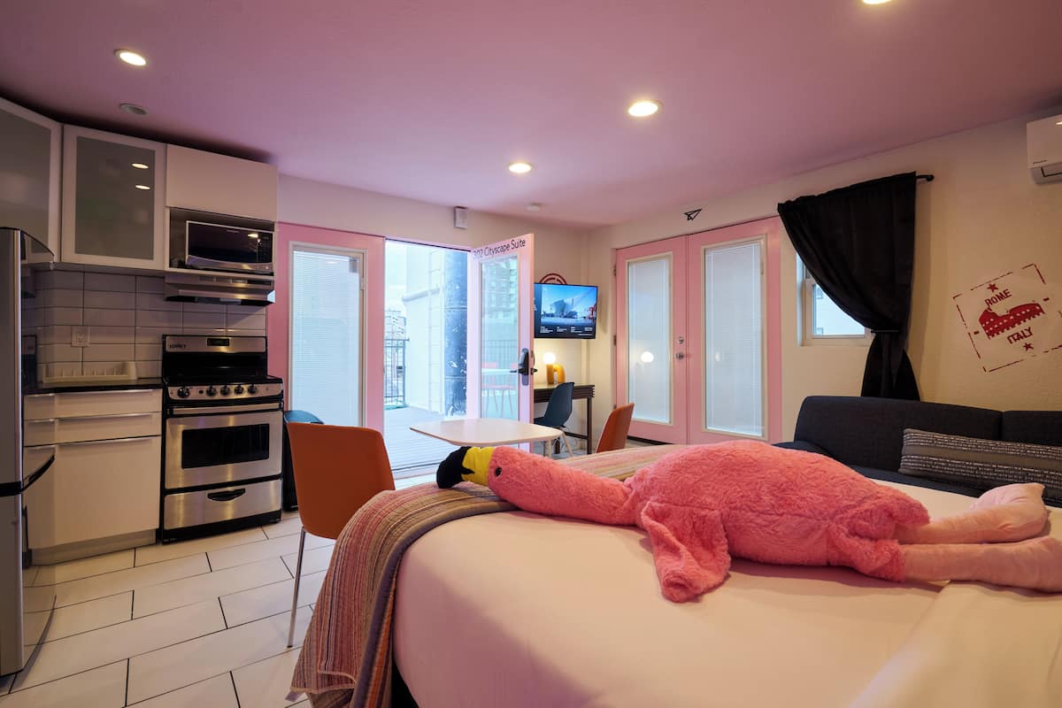 Habitación de hotel con kitchenette, una cama con manta rosa, televisor y ventana con vista a edificios.