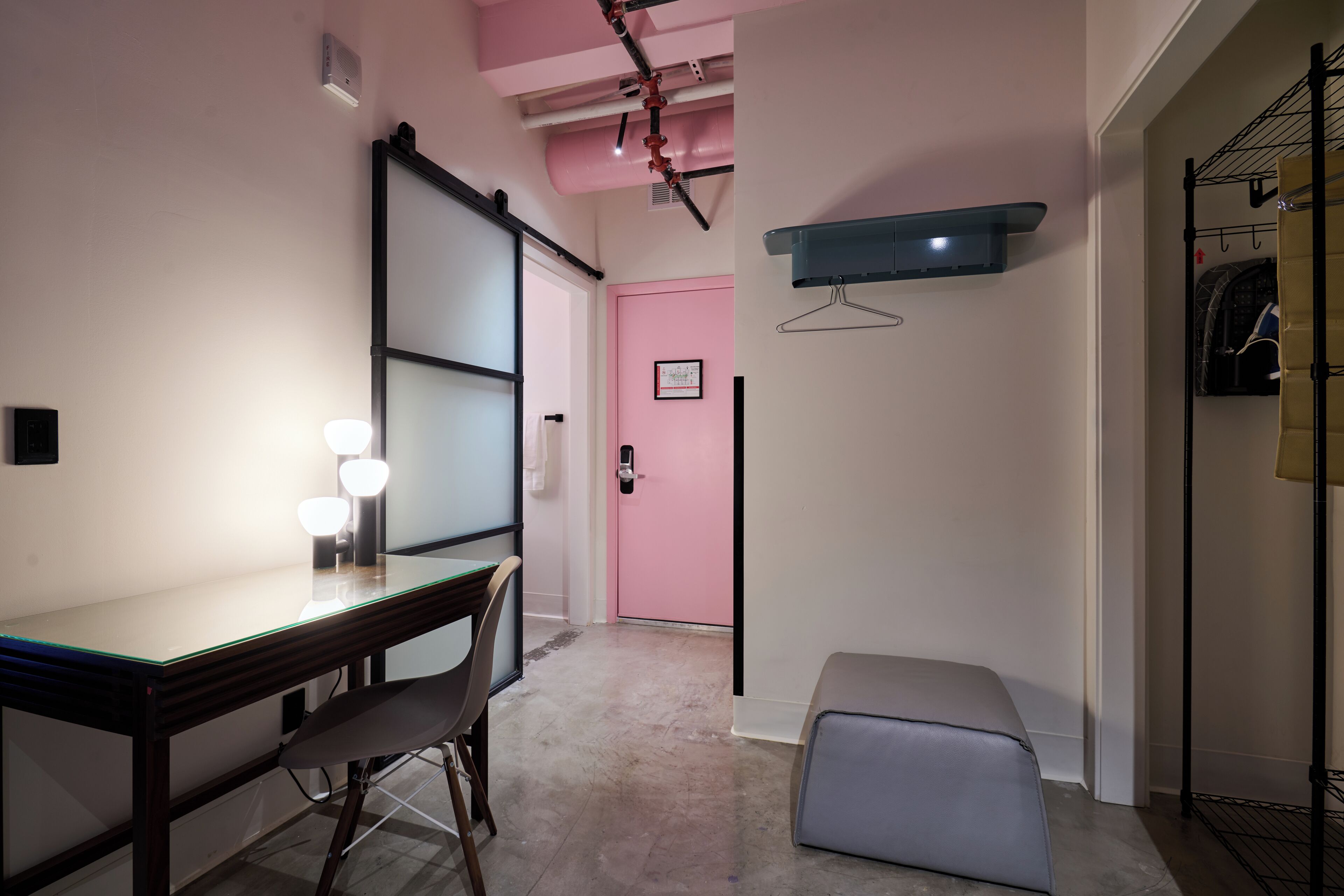 Una habitación moderna con un escritorio de vidrio, una silla, un banco, una puerta rosa y un perchero.