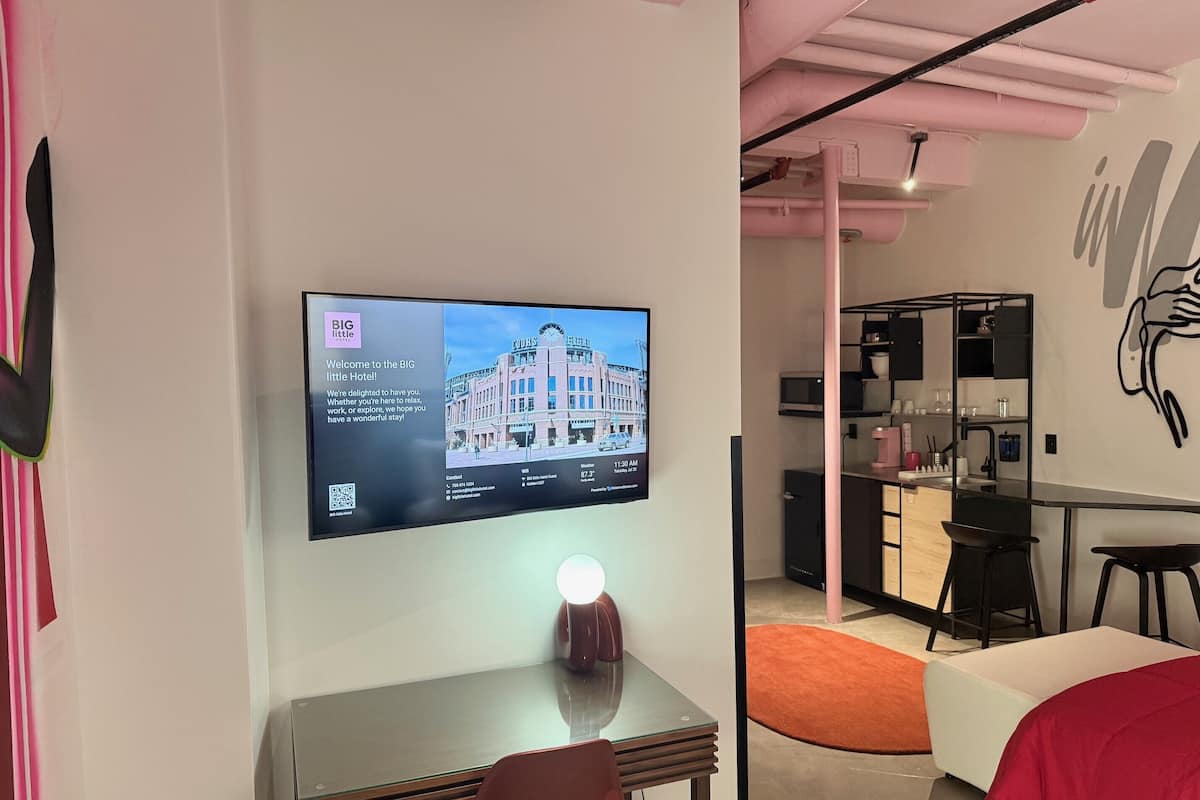 Una habitación de hotel moderna con una televisión de pantalla plana mostrando un mensaje de bienvenida, un escritorio con una silla, una cama con una colcha roja y una zona de kitchenette.