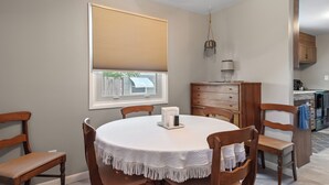 A dining area with a round table, wooden chairs, a white tablecloth, a window with blinds, and a kitchen in the background. - ⁂Pet-friendly⁂Fenced yard w/deck⁂Pool Table⁂ (Rapid City)