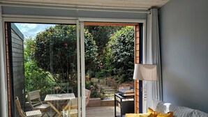 A modern living room with a glass door leading to a wooden deck, a sofa with cushions, a small table, and a lamp.