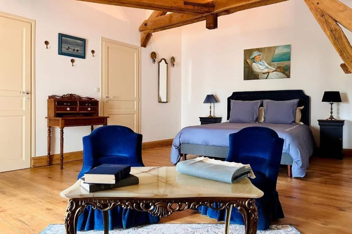 A bedroom with a bed, two chairs, a table, a dresser, and a painting on the wall.