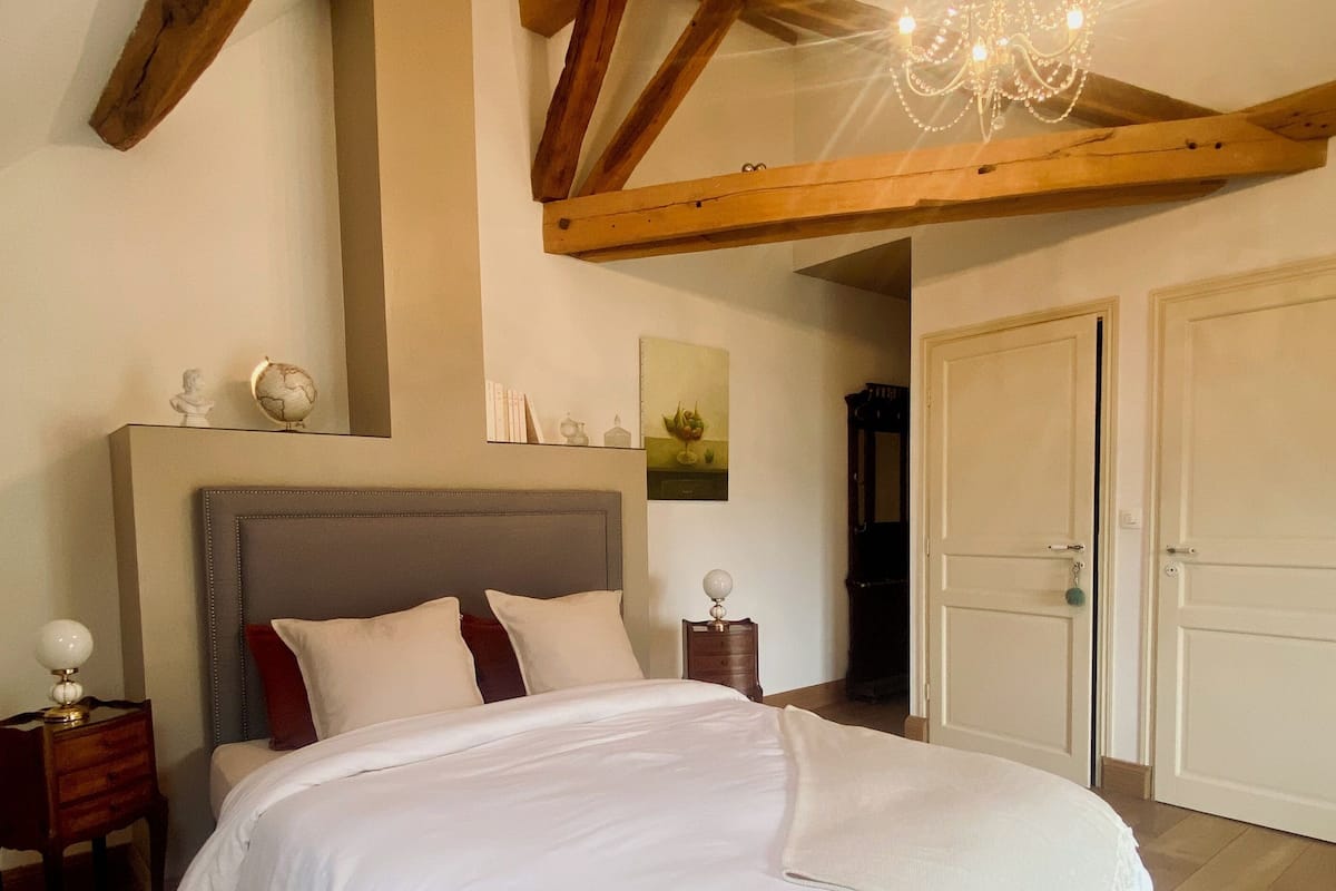 A bedroom with a large bed, wooden beams, a chandelier, and a door.