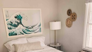 A neatly made bed with a white quilt, a wall-mounted artwork of a wave, a bedside lamp, and a small round table with a lamp. - Digs 3 of 5: 2BDRM, Open & Airy, Private Parking, Central, 2nd Floor & More! (Evansville)