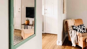A room with a green-framed mirror, a television, a sofa with a patterned throw, and a wooden floor. - Digs 3 of 5: 2BDRM, Open & Airy, Private Parking, Central, 2nd Floor & More! (Evansville)