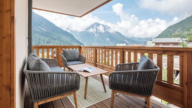 A balcony with wicker chairs and a table, overlooking a mountainous landscape.