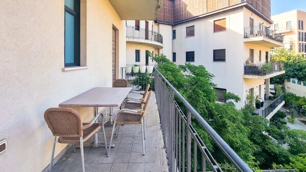 Un balcon doté d’une table et de chaises, entouré d’une rambarde en métal, donnant sur une cour verdoyante et sur les bâtiments adjacents.
