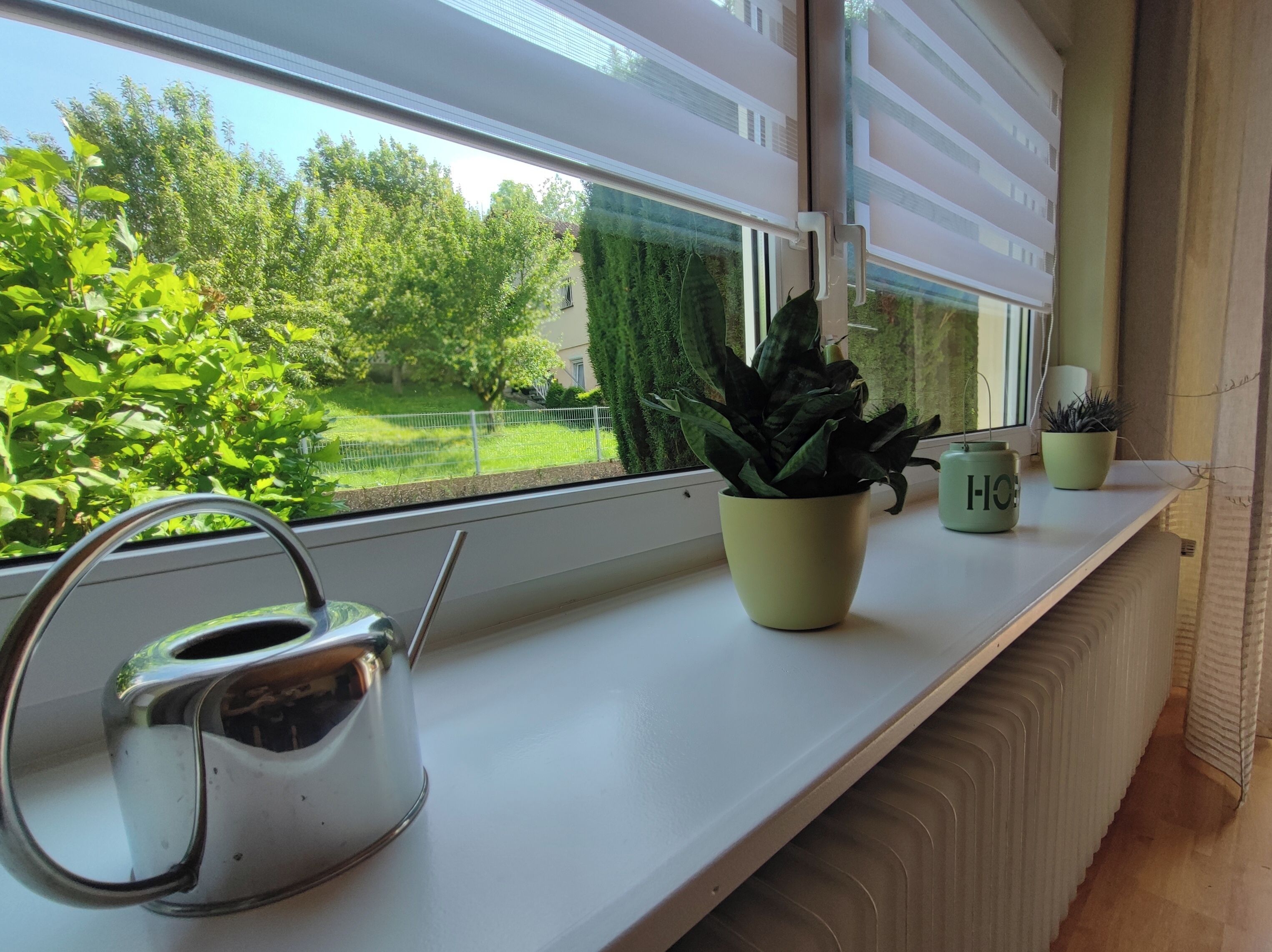 Ein Fenster mit Blick auf einen grünen Garten, ein Wasserkocher und zwei Blumentöpfe auf einer weißen Ablage.