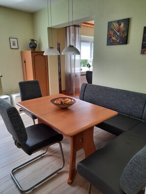 A dining area with a wooden table, black cushioned chairs, a bowl of fruit, a sofa, and a painting on the wall.