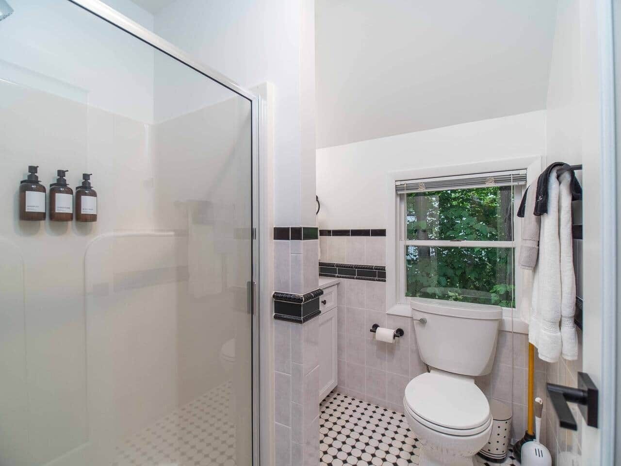 A bathroom with a glass shower enclosure, a toilet, a window, and toiletries on the wall.