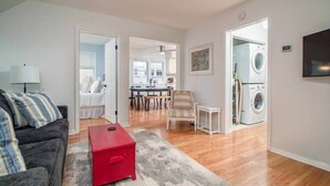 Un salon avec un canapé gris, un pouf rouge et un tapis à motifs. On y trouve également un coin repas avec une table et des chaises, et une cuisine visible à travers une porte ouverte.
