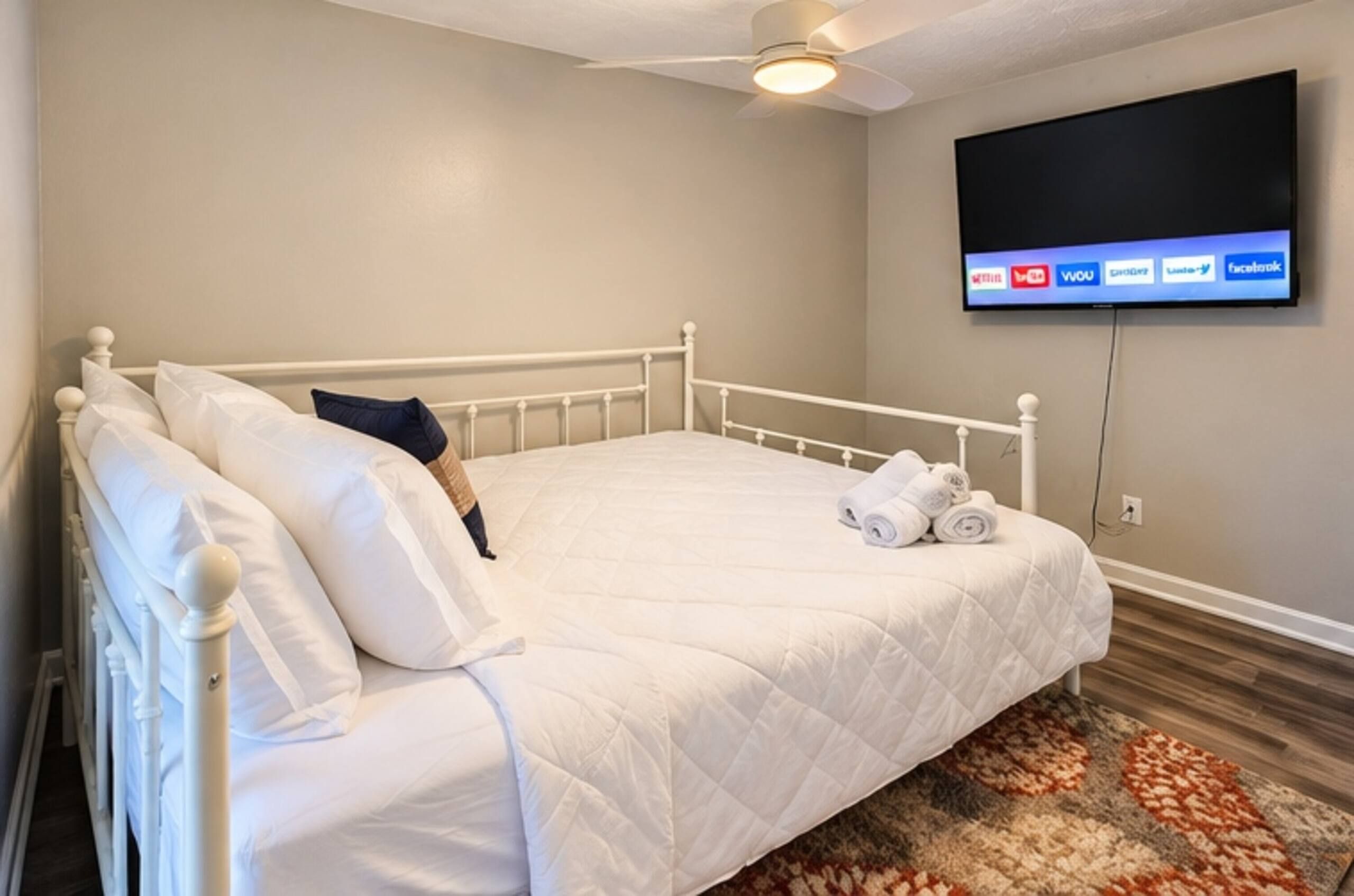 Een slaapkamer met een bed, kussens, een plafondventilator, een aan de muur bevestigde televisie en een kamerbreed tapijt.