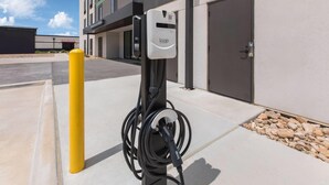 An electric vehicle charging station with a cable connected to it, a yellow bollard, and a building in the background.