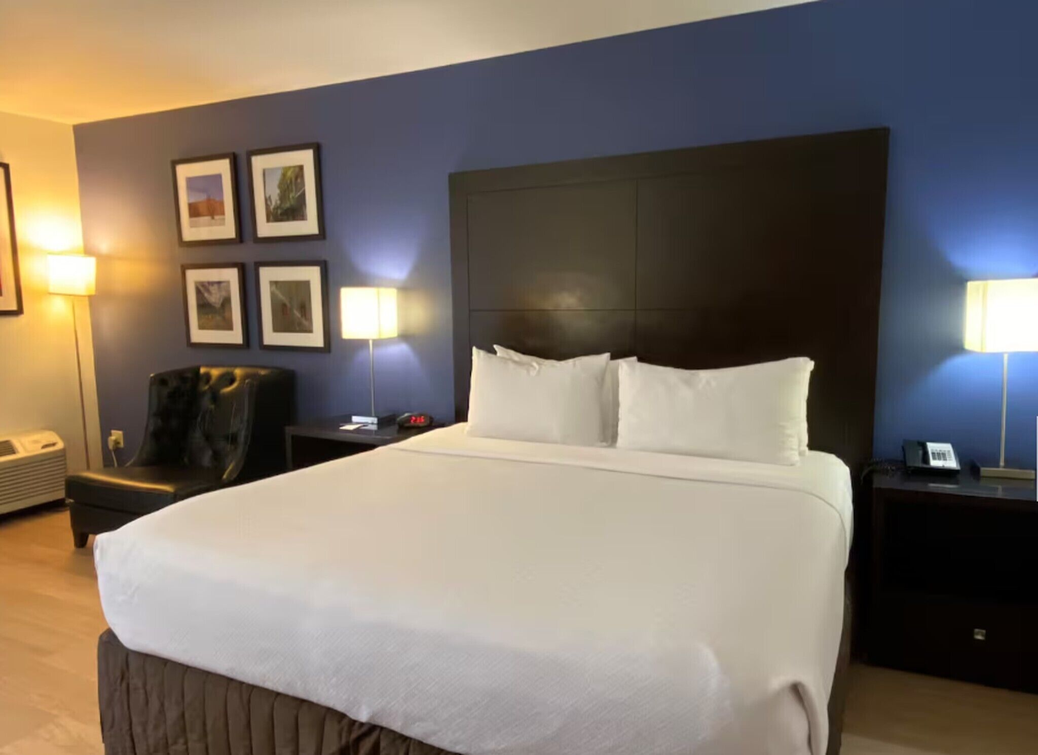 Ein Hotelzimmer mit einem großen Bett, einem Kopfteil, einem Sessel, einem Nachttisch, einer Lampe und gerahmten Bildern an der Wand.