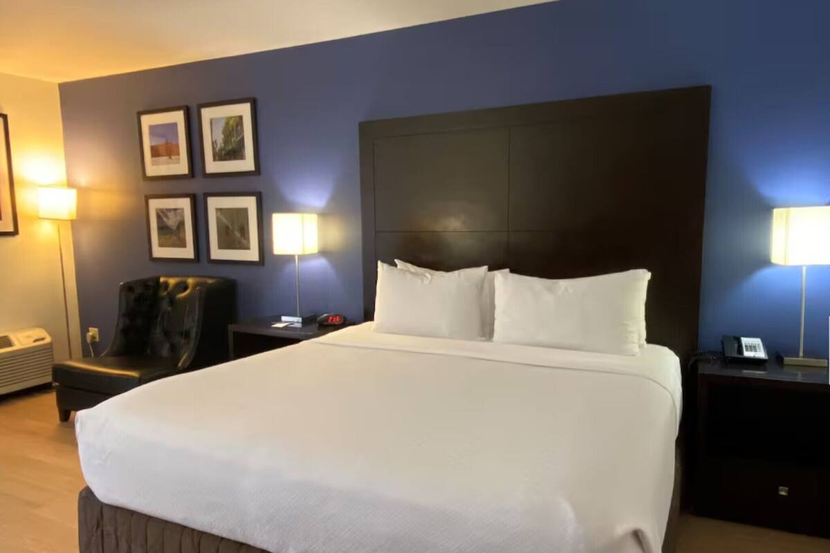 A hotel room with a large bed, a headboard, a chair, a nightstand, a lamp, and framed pictures on the wall.