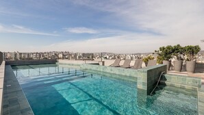 Piscina infinita en la azotea con vistas a la ciudad, sillones de descanso y plantas en macetas.