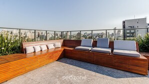 Zona de estar en la azotea con banca de madera y asientos acolchados, con vistas a un paisaje urbano.