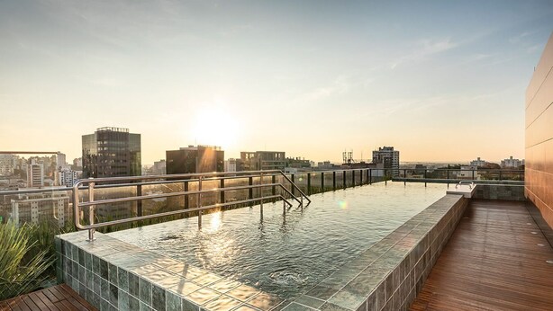 Una piscina infinita en la azotea con vista a la ciudad, una terraza de madera y una baranda metálica.