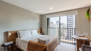 A modern hotel room with a bed, a small table with wine glasses, a balcony with a view of buildings, and a plant on the wall. - High-standard Studio in Moinhos Gos1007 (Porto Alegre)