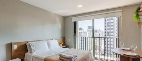 Habitación de hotel moderna con cama, una mesita con copas de vino, balcón con vistas a edificios y una planta en la pared.