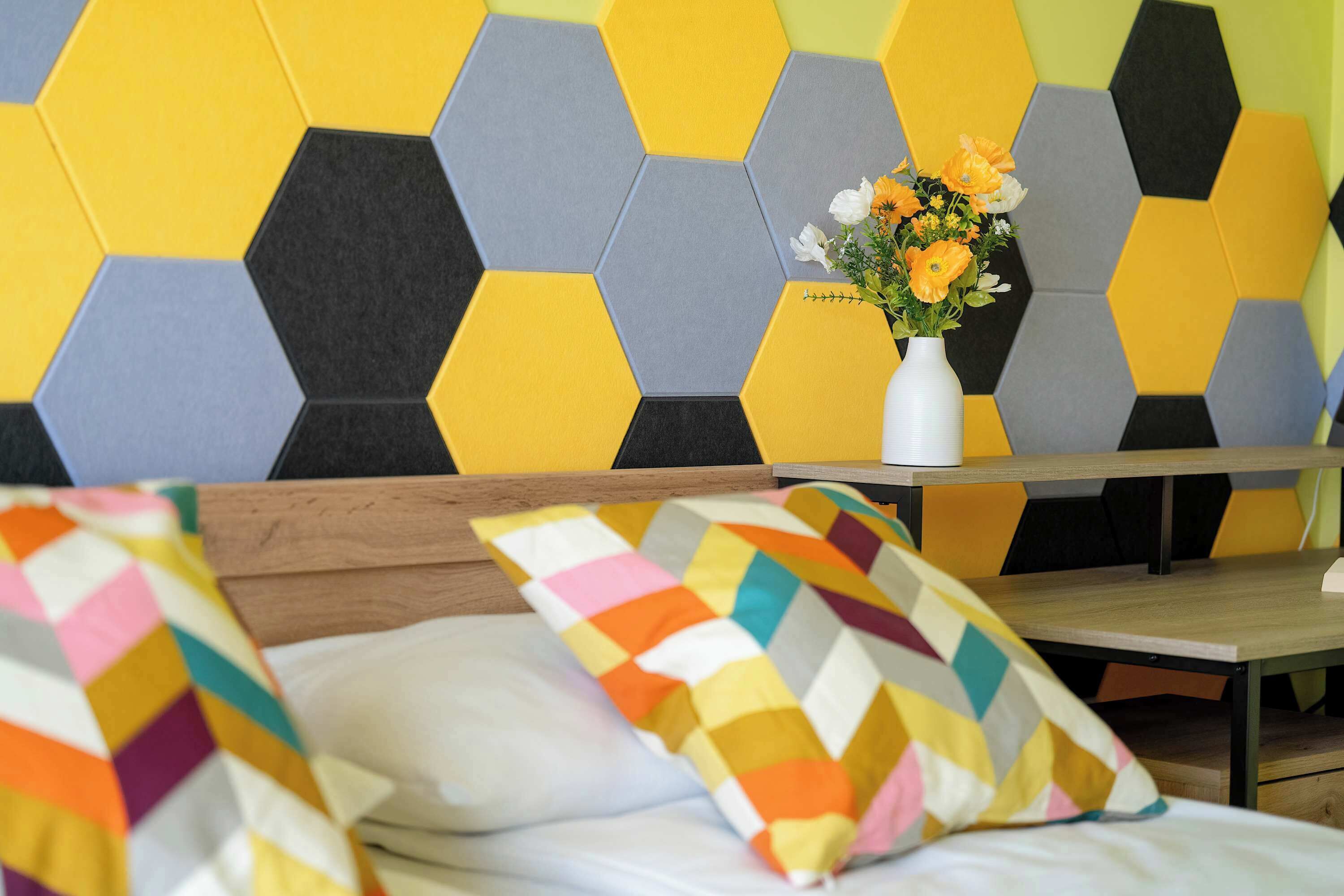 A hotel room with a bed, a wooden headboard, a table with a vase of flowers, and a wall with a geometric pattern.