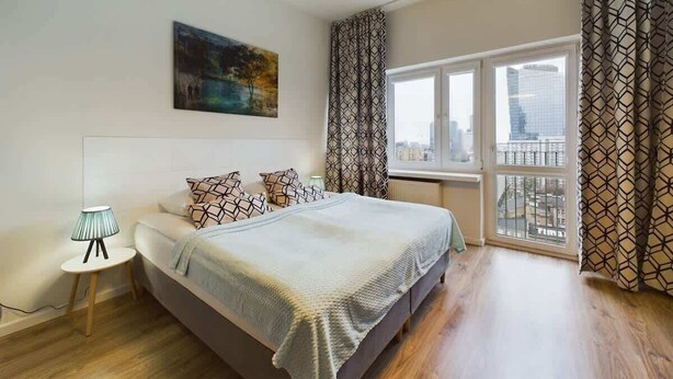 Un dormitorio moderno con una cama grande, una lámpara de noche, vistas de la ciudad a través de la ventana y cortinas estampadas.