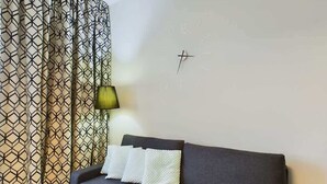 Un salón moderno con un sofá gris oscuro, tres cojines blancos, una cortina con estampado geométrico y un reloj de pared minimalista.