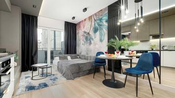 A modern living room with a sofa, a dining table with chairs, a kitchen area, and a large window with curtains.