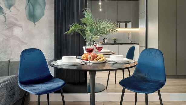 A modern dining area with a round table, two blue chairs, a potted plant, and a fruit bowl.