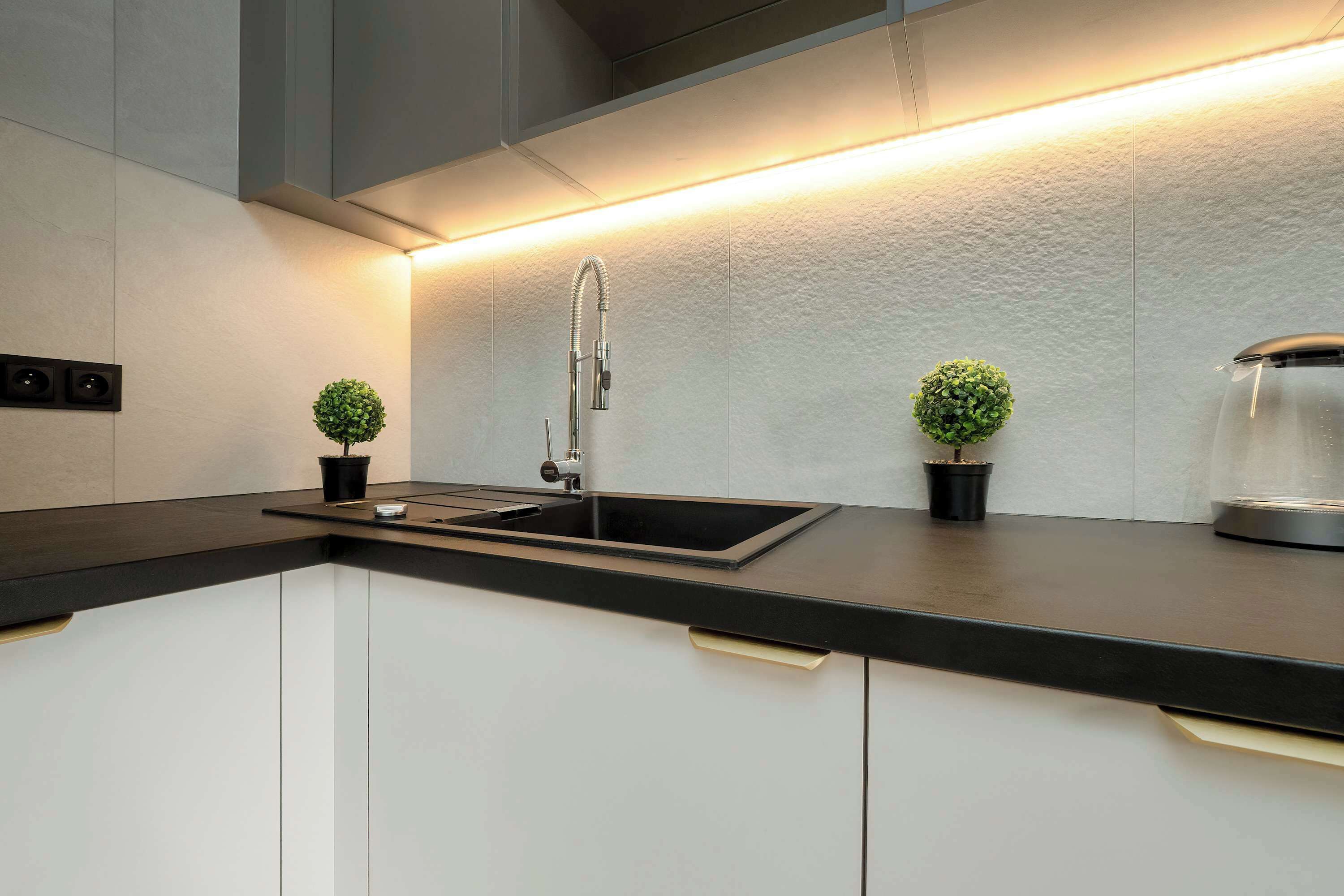 Een moderne keuken met een zwart aanrecht, witte kastjes, een roestvrijstalen spoelbak en twee kleine potplanten.