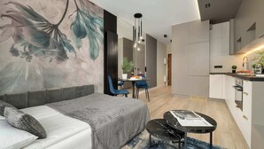 A modern hotel room with a sofa, a dining table with chairs, a small kitchen area, and a large floral mural on the wall.