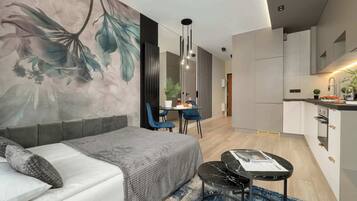 A modern hotel room with a sofa, a dining table with chairs, a small kitchen area, and a large floral mural on the wall.